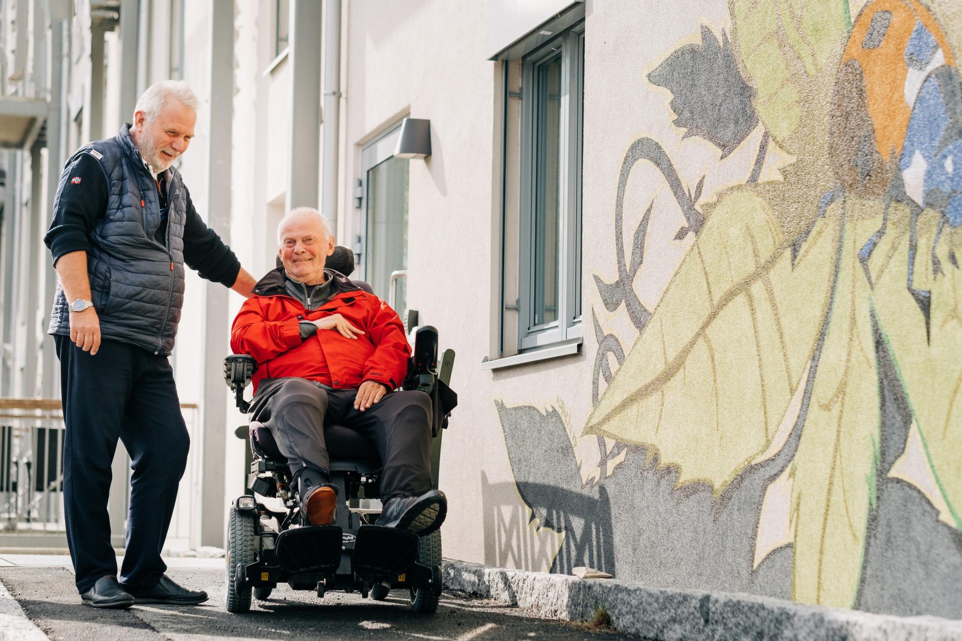To menn er utendørs på et fortau, den ene i en motorisert rullestol og den andre står ved siden av ham og smiler. Et stort veggmaleri er synlig på veggen ved siden av dem.